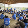 Group of people in blue T-shirts gathered in an open-air pavilion, with some sitting on the floor and one person standing and speaking.