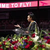 A graduation speaker in a cap and gown gestures at a podium, with flowers in the foreground and a "TIME TO FLY!" banner above.